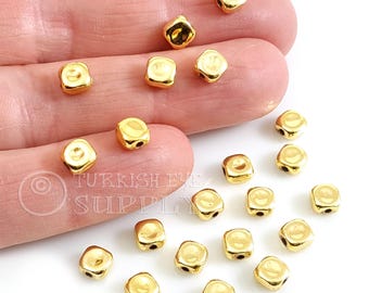 May include: A close-up of 20 gold-colored square beads with a hole in each corner. The beads are scattered on a white surface.