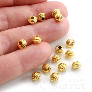 Peut inclure: Une collection de petites perles rondes de couleur dorée avec une texture nervurée horizontale. Chaque perle présente un trou central pour l'enfilage. Les perles ont une finition métallique brillante et sont éparpillées sur une surface claire.