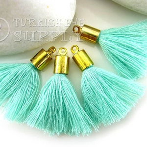 May include: Four turquoise blue tassel charms with gold metal caps. The tassels are made of soft, fluffy yarn and are perfect for adding a touch of bohemian style to jewelry, bags, or home decor.