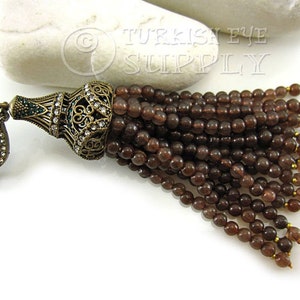 May include: A brown beaded tassel with a decorative metal top featuring clear rhinestones and green accents. The tassel has a metal loop for attaching to a necklace or bracelet.