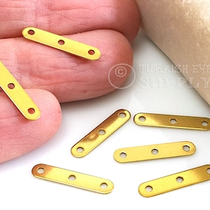 May include: Seven gold-colored metal connector pieces with three holes each. The pieces are rectangular and have rounded edges. The pieces are arranged on a white surface.