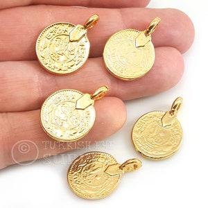 May include: Five gold-colored metal coin charms with intricate designs and a loop for hanging. The charms are on a hand.