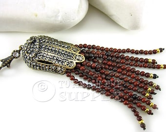 May include: A gold-toned Hamsa hand charm with clear crystals, attached to a tassel of red jasper beads.