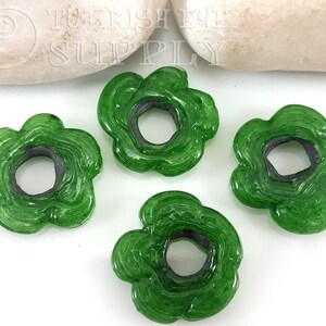 May include: Four green glass flower-shaped beads with a hole in the center. The beads have a swirl pattern and a dark green rim.