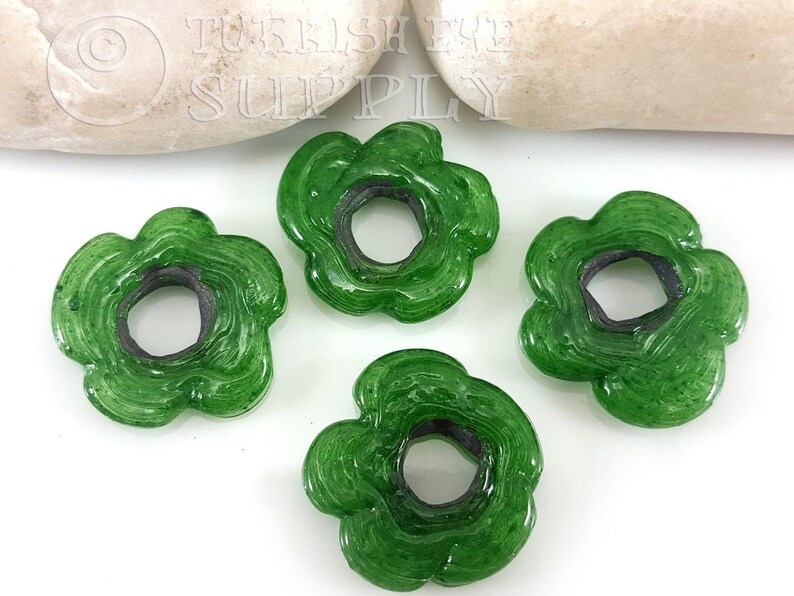 May include: Four green glass flower-shaped beads with a hole in the center. The beads have a swirl pattern and a dark green rim.