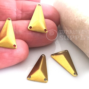 Puede incluir: Cuatro abalorios de metal de forma triangular de color dorado con tres agujeros cada uno. Los abalorios están sobre una superficie blanca.