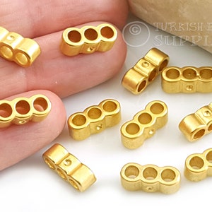 May include: Ten gold-coloured metal spacer beads with three holes each. The beads are arranged on a hand and a light brown surface. The text "TURKISH EYE SUPPLY" is visible in the background.