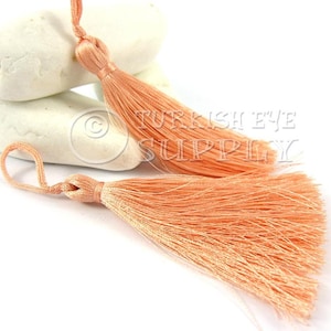 May include: Two peach-colored tassels with a long, thin cord. The tassels are made of soft, silky fibers and are tied together with a knot.