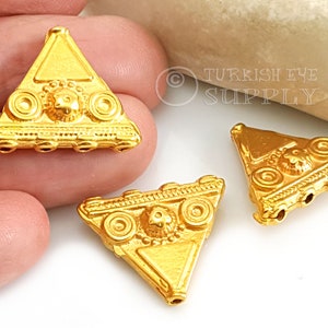 May include: Three gold-tone triangle-shaped pendant charms with intricate details and a raised design. Each charm has a circular pattern and a raised centre design. The charms have holes for stringing.