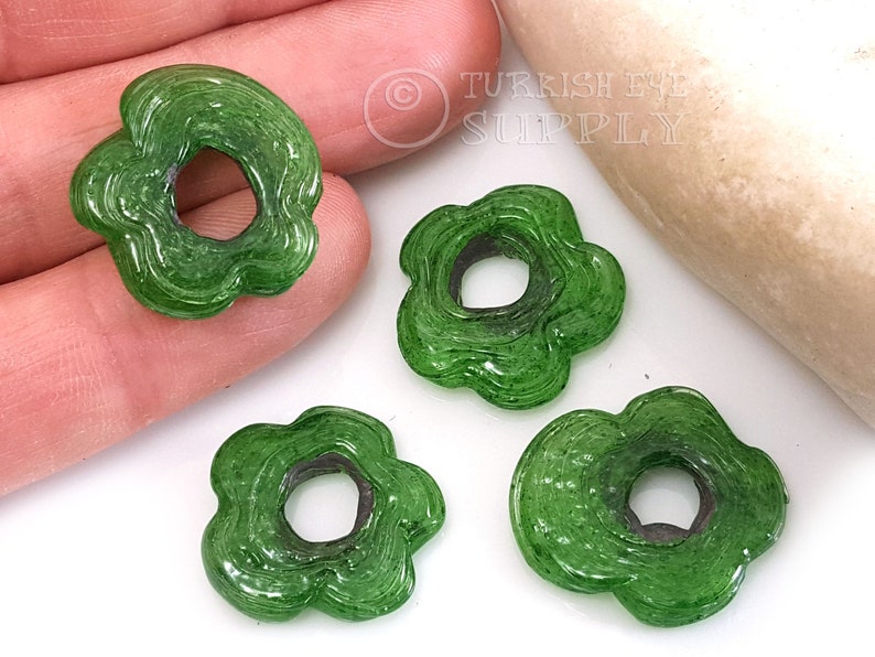 May include: Four green glass flower-shaped beads with a hole in the center. The beads have a textured, swirled design.