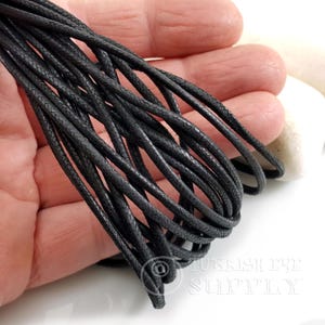 May include: Close-up of a bundle of black waxed cotton cord. The cord is tightly woven and appears to be durable. The cords are held in a hand, with a white background.