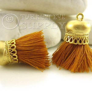 May include: Two gold tassel tops with a decorative filigree design and a mustard yellow tassel.  The tassel tops are on a white background.