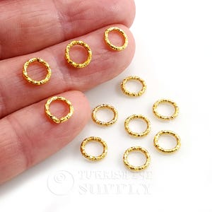 May include: A collection of small, circular gold-coloured jump rings with a textured, hammered surface. These metal rings feature a small opening, allowing them to be used as connectors for jewellery making. The rings are arranged in a scattered pattern.