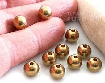 May include: A close-up of ten gold-colored metal beads. The beads are round and have a hole in the center. The beads are scattered on a white surface.