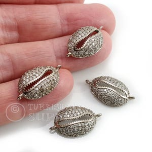 May include: Four silver-toned, oval-shaped jewelry components. Each component is encrusted with small, clear stones and features a central, open design. The components have small loops on each end for attachment.