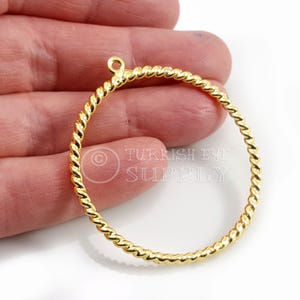 Puede incluir: Un componente de pendiente de aro dorado, estilo cuerda trenzada. El dije circular tiene un pequeño lazo en la parte superior para la fijación. El texto "TURKISH EYE SUPPLY" es visible en el fondo.