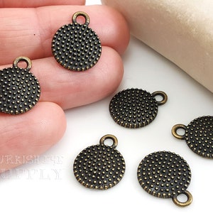 以下が含まれることがあります： ブロンズ色の丸いチャームで、テクスチャード加工された点状の表面と上部に小さなループが付いています。 チャームは小さく、おそらくジュエリー作りや工芸品用です。 画像にはいくつかのチャームが写っています。