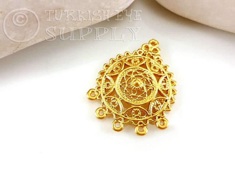 May include: Gold-toned metal pendant with a circular filigree design and four hanging loops. Turkish Eye Supply is printed on the background.