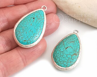 Op de afbeelding: Twee turquoise blauwe traanvormige hangers met zilverkleurige metalen instellingen. De hangers hebben een gebarsten textuur en zijn perfect voor het maken van sieraden.
