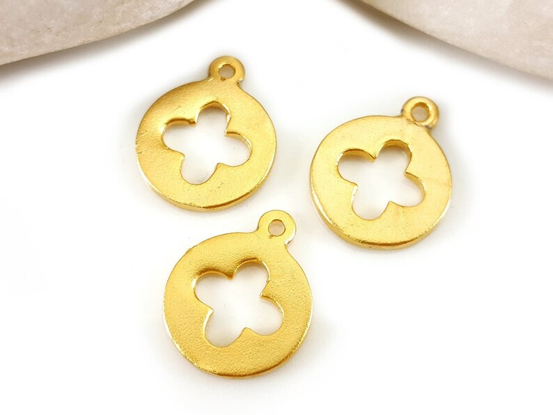 May include: Three gold-colored metal charms with a four-leaf clover cutout design. The charms are round and have a loop at the top for attaching to jewelry or other crafts.