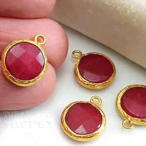 May include: Four gold-toned pendants with red gemstone cabochons. Each pendant has a loop for attaching to a chain or cord. The pendants are arranged on a white surface.