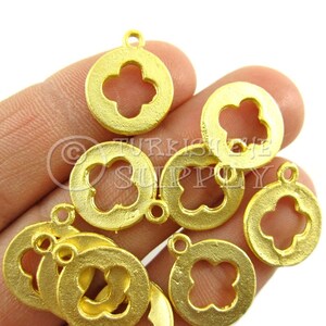 May include: A close-up of a hand holding a pile of gold-colored metal charms. Each charm is a circle with a four-leaf clover cutout in the center. The charms have a slightly textured surface.