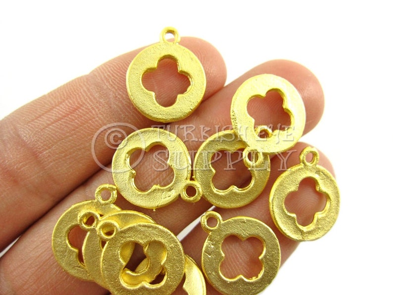 May include: A close-up of a hand holding a pile of gold-colored metal charms. Each charm is a circle with a four-leaf clover cutout in the center. The charms have a slightly textured surface.