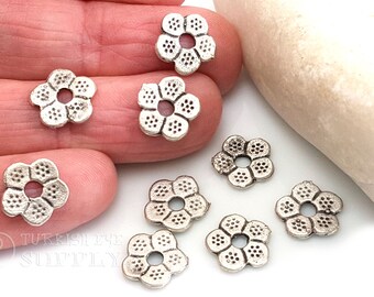 May include: Silver flower-shaped metal beads with a hole in the center. The beads are about 1 cm in diameter and have a textured surface.