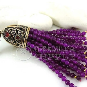 Könnte beinhalten: Lilafarbene Amethyst-Edelsteinkugeln mit einem goldenen Quastenkopf. Der Quastenkopf hat einen roten Stein und komplizierte Details.  Turkish Eye Supply ist auf dem Bild gedruckt.
