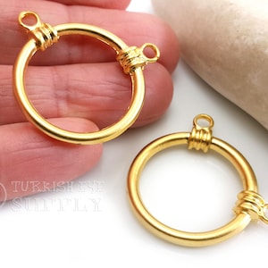 May include: Two gold-coloured, circular jewellery clasps. Each clasp has a ring shape with a small loop at the top and a decorative wrapped section. The clasps are shown against a white background.