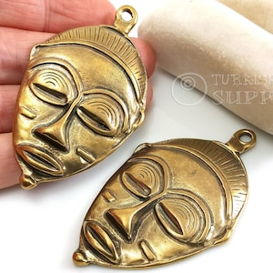 May include: Two gold-toned metal pendants shaped like African masks. The masks have detailed carvings and a raised lip around the edges.