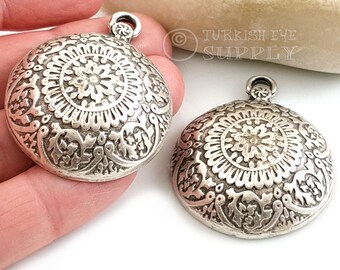May include: Two silver-toned metal pendants with intricate floral designs. The pendants are round and have a loop at the top for attaching to a chain or cord.