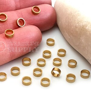 Puede incluir: Primer plano de 20 anillos de salto dorados, de aproximadamente 6 mm de diámetro, esparcidos sobre una superficie blanca. Los anillos de salto se utilizan para la elaboración de joyas y manualidades.