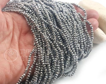 May include: Strands of small, faceted, metallic gray beads. The beads are strung together, creating multiple lines of the same material. The beads are held in a hand, with a white background and a light-colored stone visible.