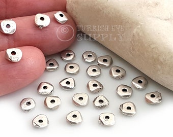 May include: A close-up of a group of silver-toned metal beads with a hole in the center. The beads are irregularly shaped and have a rough, textured surface.  The beads are scattered on a white surface.