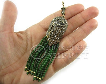 May include: A gold Hamsa hand charm with clear rhinestones and a green beaded tassel. The charm is on a hand, and the text "TURKISH EYE SUPPLY" is visible in the background.