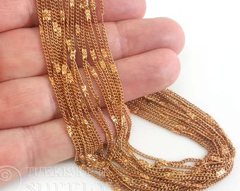 May include: Close-up of multiple strands of delicate gold-colored chain. The chains have a textured, intricate design with small floral or geometric accents. The chains are held in a hand against a white background, showcasing the jewelry's details.
