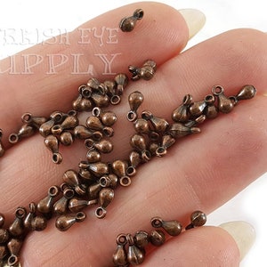 May include: A pile of small, copper-colored teardrop-shaped metal charms with a loop at the top. The charms are on a hand.