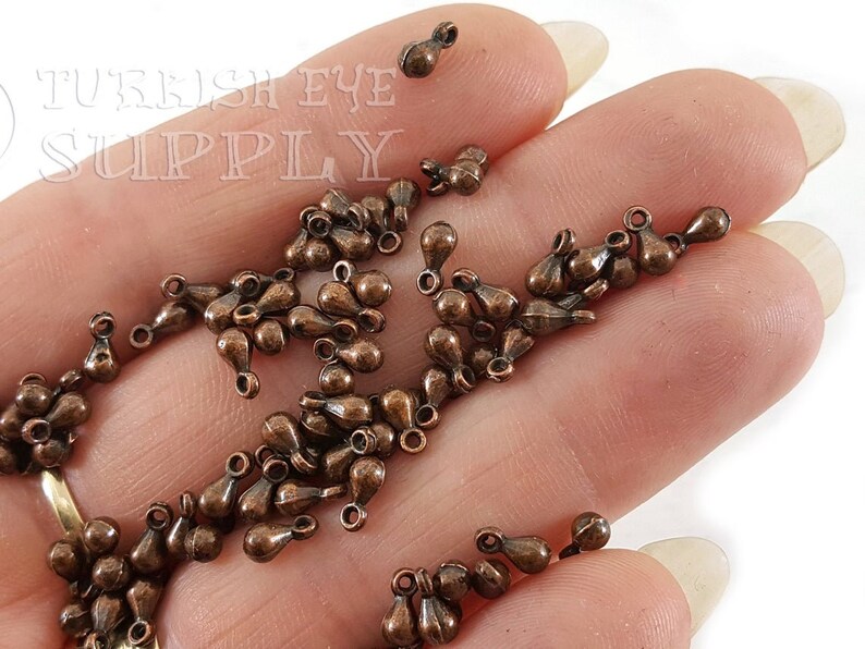 May include: A pile of small, copper-colored teardrop-shaped metal charms with a loop at the top. The charms are on a hand.