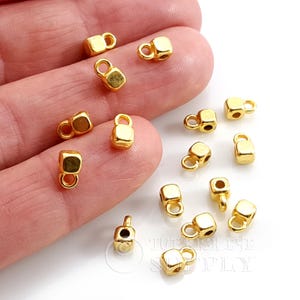 Könnte beinhalten: Eine Sammlung kleiner, goldfarbener Metallwürfelperlen, die jeweils mit einer kleinen Öse an der Oberseite versehen sind. Diese Metallkomponenten sind für die Schmuckherstellung gedacht und liegen verstreut im Bild, mit einer glatten Oberfläche.