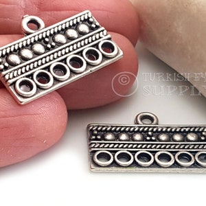 Puede incluir: Dos colgantes de metal de tono plateado con un diseño decorativo que presenta una fila de cuentas, un borde de cuerda y cinco agujeros redondos. Los colgantes tienen un bucle en la parte superior para sujetarlos a un collar o pulsera.