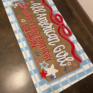 May include: A rectangular sign with a brown background and a blue and white checkered border. The sign reads "All American Girl" in white script with a red ribbon design. It also says "Happy First Birthday Callahan" with star and flag accents.