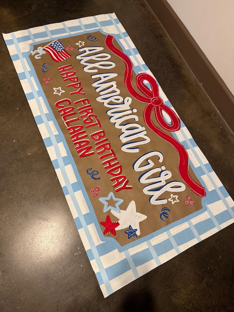 May include: A rectangular sign with a brown background and a blue and white checkered border. The sign reads "All American Girl" in white script with a red ribbon design. It also says "Happy First Birthday Callahan" with star and flag accents.