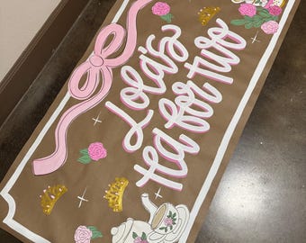 May include: A brown paper banner with the text "Lola's Tea for Two" in pink and white script. The banner features a pink bow, roses, crowns, and teacups with floral designs. The border is white.