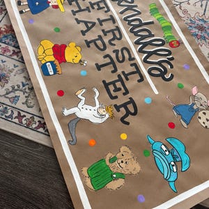 May include: A brown paper banner with white trim, featuring colorful illustrations of storybook characters. The banner reads "Kendall's FIRST CHAPTER" in black script. Winnie the Pooh, a caterpillar, and other characters are depicted.