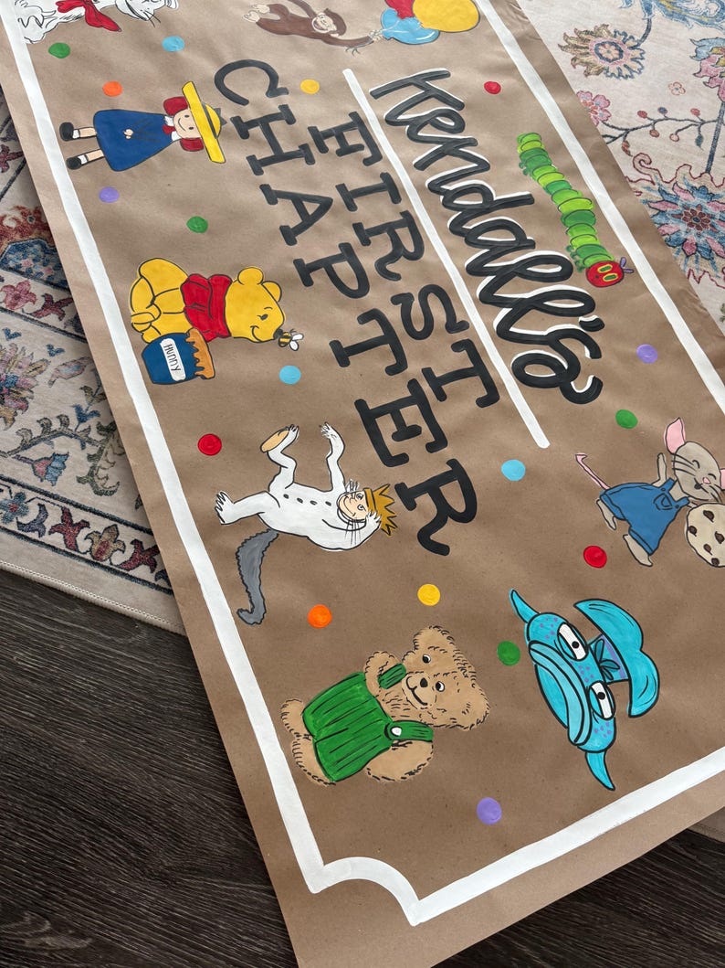 May include: A brown paper banner with white trim, featuring colorful illustrations of storybook characters. The banner reads "Kendall's FIRST CHAPTER" in black script. Winnie the Pooh, a caterpillar, and other characters are depicted.
