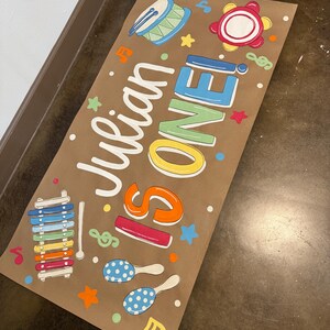 May include: A brown paper banner with the text "Julian IS ONE!" in white, blue, orange, and red. The banner is decorated with colorful musical instruments, stars, and musical notes. A drum, tambourine, xylophone, and maracas are featured.