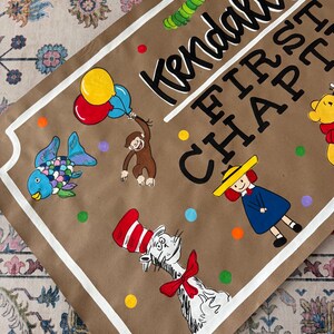 May include: A brown paper sign with white trim and the text "Kendall's FIRST CHAPTER". The sign features colorful illustrations of a fish, monkey, Cat in the Hat, and other cartoon characters. Balloons and polka dots are also included.