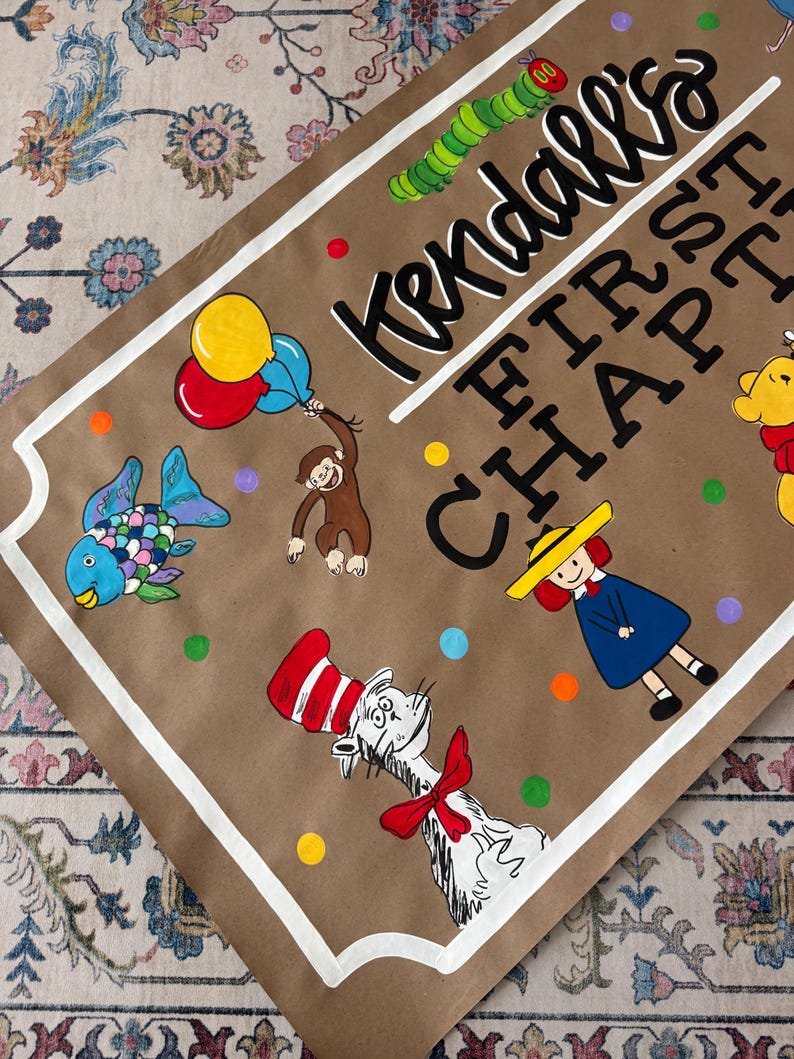 May include: A brown paper sign with white trim and the text "Kendall's FIRST CHAPTER". The sign features colorful illustrations of a fish, monkey, Cat in the Hat, and other cartoon characters. Balloons and polka dots are also included.