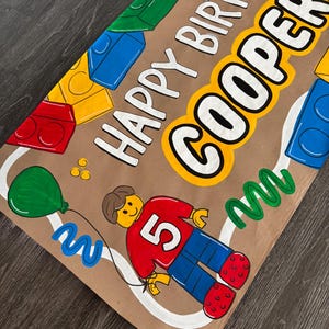 Pu&ograve; includere: Banner di compleanno dipinto a mano su carta marrone. Presenta il testo "HAPPY BIRTHAY COOPER!" in bianco con un contorno giallo. Include illustrazioni colorate di blocchi giocattolo, un palloncino e una figura con il numero 5.